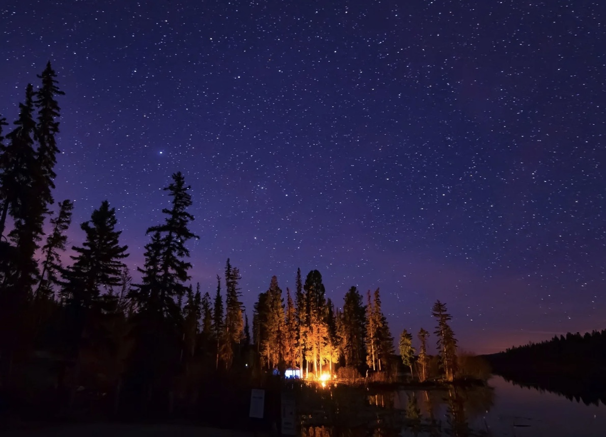 A starry night sky over a cabin lit warm beside a calm lake